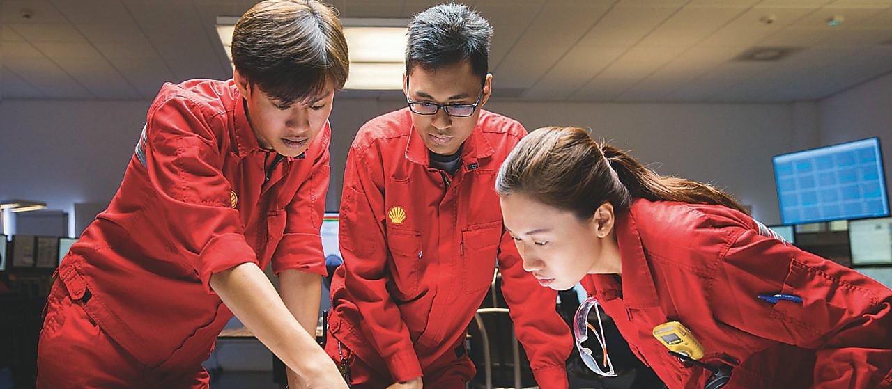 Dos hombres y una mujer, con uniformes de seguridad rojos, discuten en equipo en una oficina de Shell.