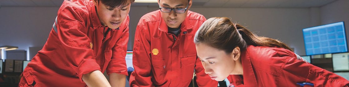 Dos hombres y una mujer, con uniformes de seguridad rojos, discuten en equipo en una oficina de Shell.