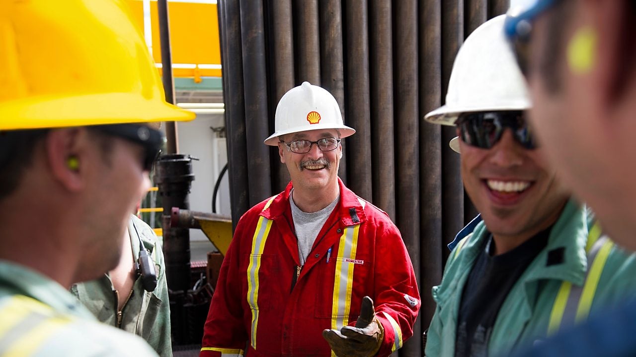 Un grupo de hombres, trabajando en una planta y vestidos con uniformes de seguridad, cascos y lentes, se sonríen entre sí.