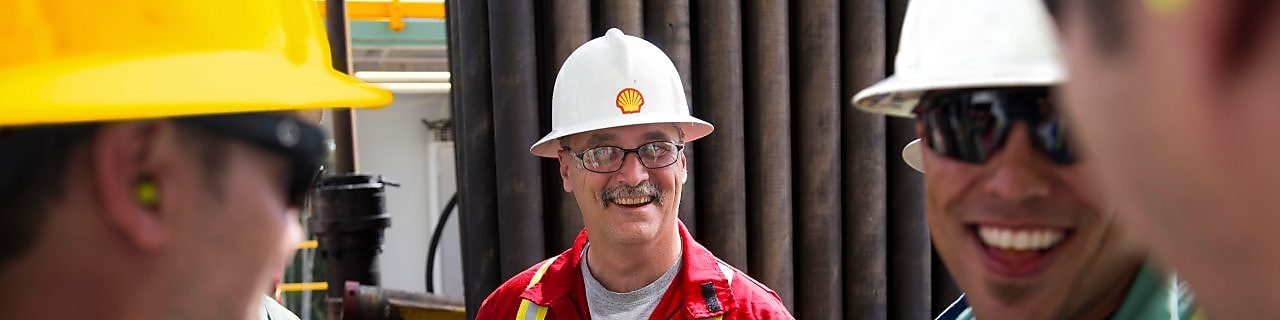 Un grupo de hombres, trabajando en una planta y vestidos con uniformes de seguridad, cascos y lentes, se sonríen entre sí.