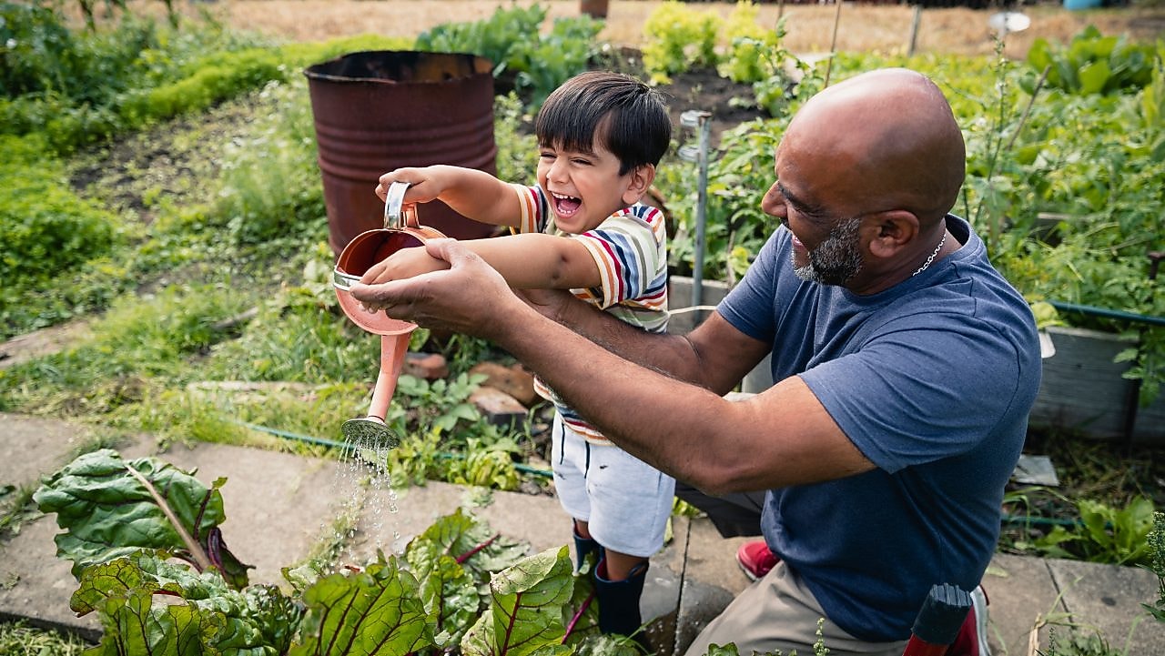 Hombre ayudando a un niño sonriente a regar plantas en un jardín