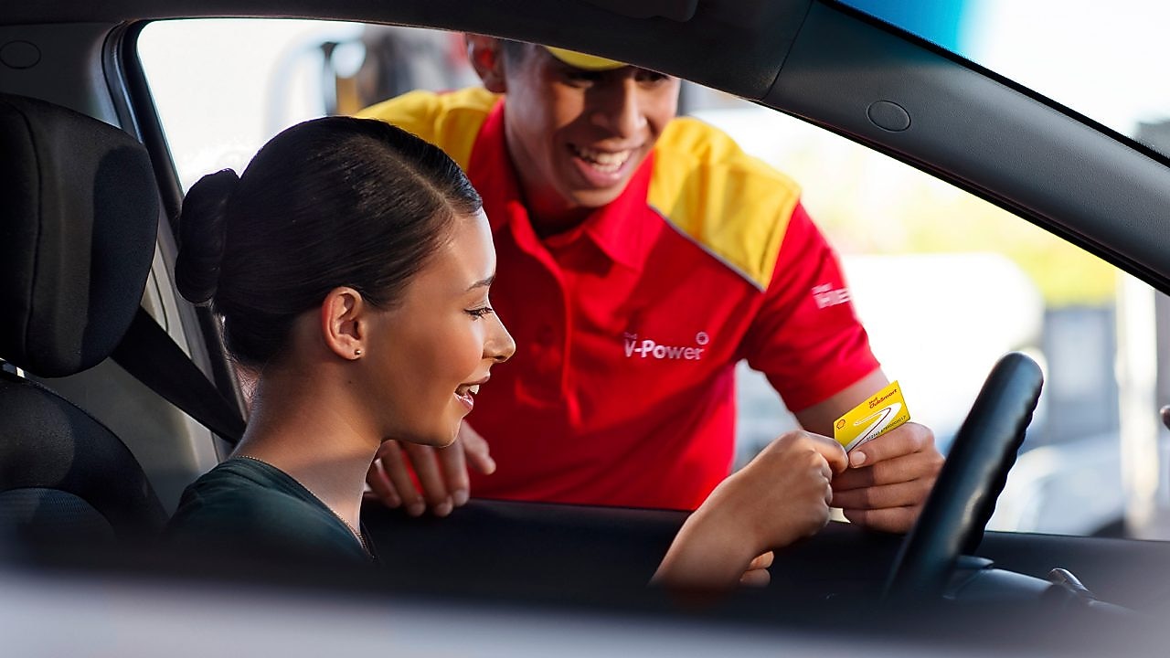 Mujer al volante, en un coche parado, siendo atendida por un empleado de una gasolinera Shell. Tiene el pelo negro y sonríe, lleva uniforme y una gorra roja. Ella le enseña una tarjeta Shell.